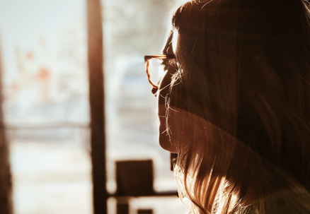 A woman with glasses gazes out a sunlit window, possibly contemplating financial matters