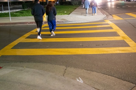 Night-time crosswalk shot showing two people walking along a brightly painted yellow crosswalk