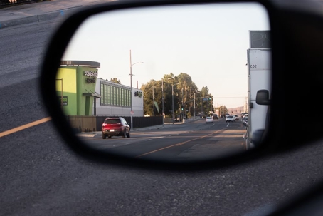 Car side mirror view shows a street with a car, building, and greenery in the background