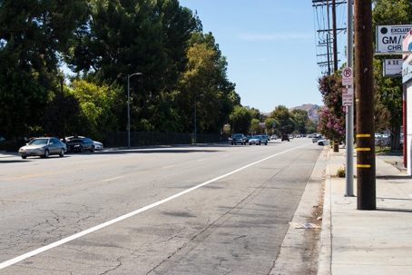 A sunlit street is lined with parked cars and trees